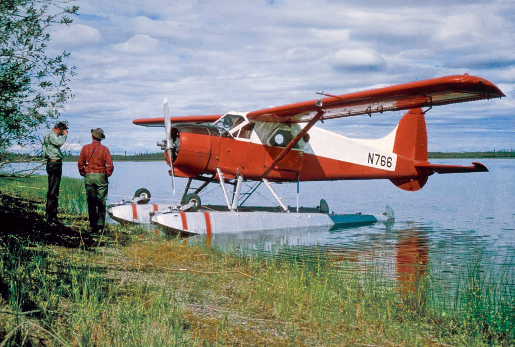 Two people standing on a shore next to a de Havilland Beaver.