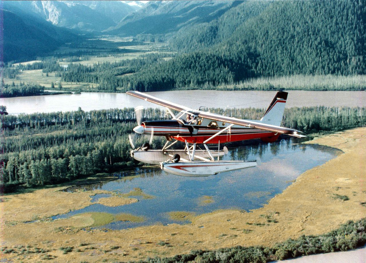 The improved Mk.3 Turbo Beaver aircraft flying over a body of water. Trees and mountains in the backgrund.