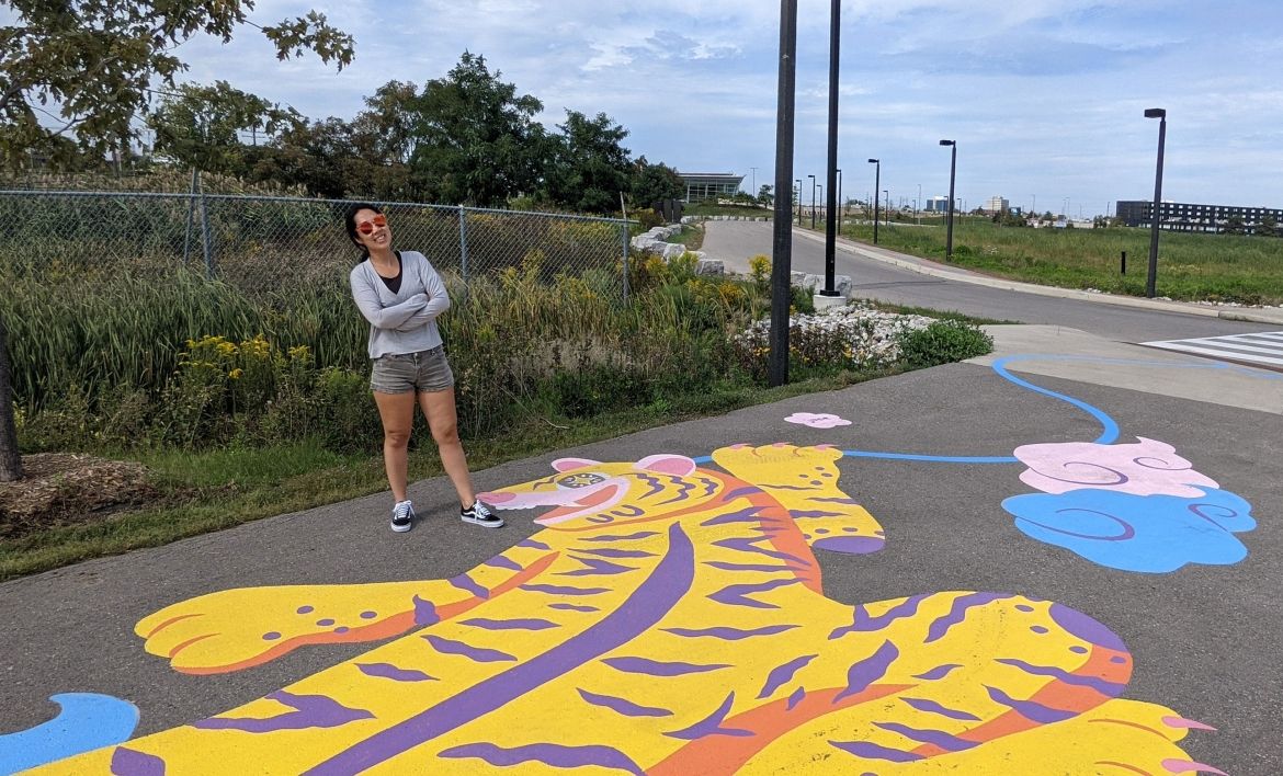 Artist Jieun June Kim standing proudly over her painting of a bright yellow tiger on a paved path.