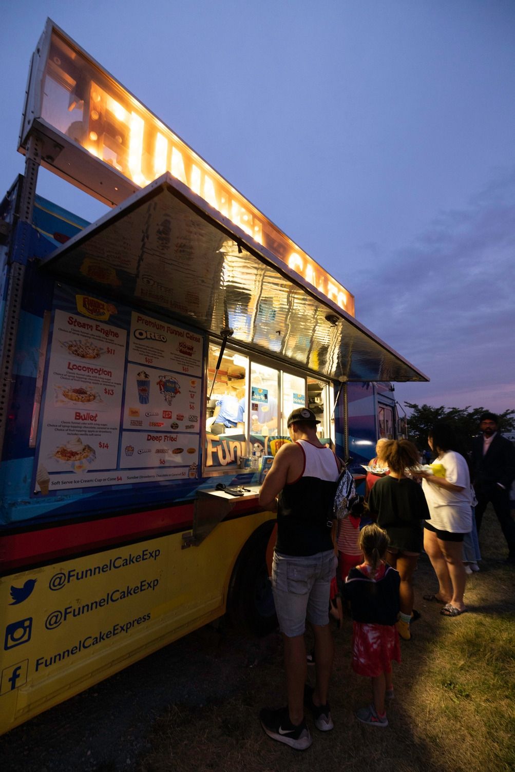 A funnel cake food truck illuminated against the night with customers in front