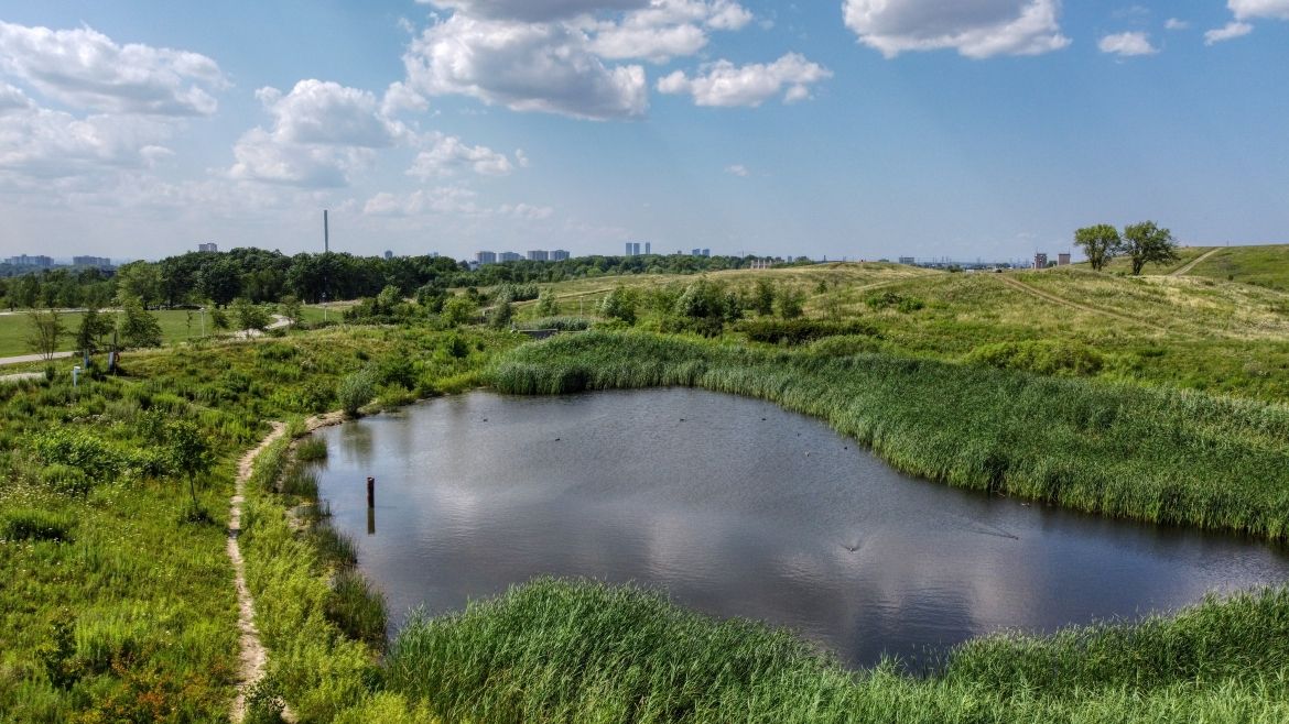 An aerial shot of a small pond, surrounded by green grass and hills.