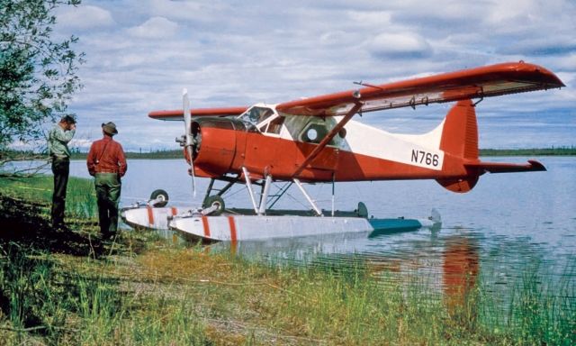 Two people standing on a shore next to a de Havilland Beaver.