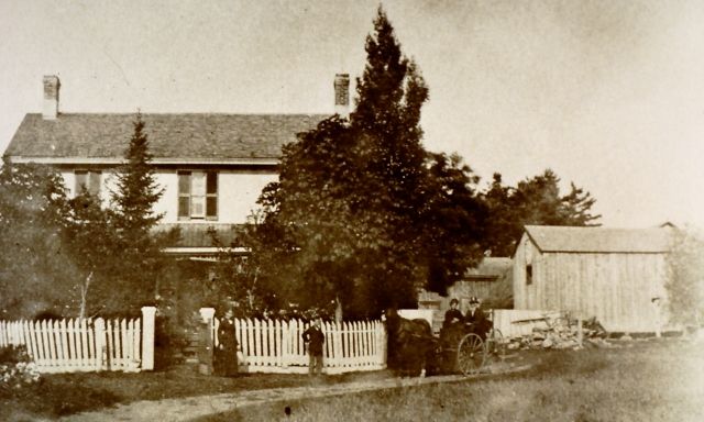 A black and white photo of a house with a white picket fence. A couple in a horse-drawn carriage are out front.