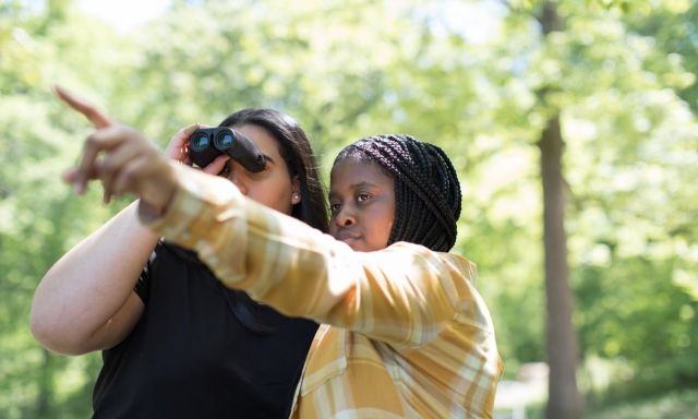 Two girls looking through binoculars in a forest.