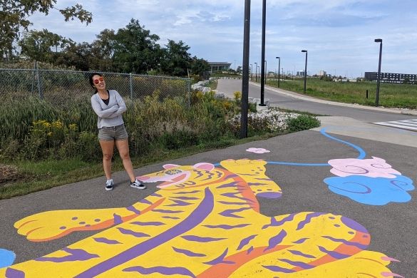 Artist Jieun June Kim standing proudly over her painting of a bright yellow tiger on a paved path.